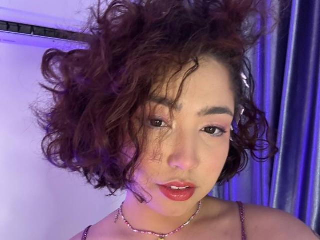 StarlitGaze has curly brown hair, red lipstick, and a pink hairclip. She's wearing a pink strap top with a pink necklace, looking slightly to the side.