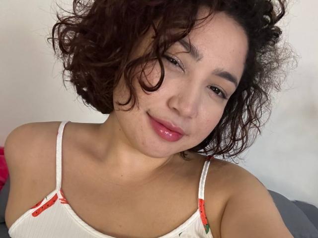 StarlitGaze has curly brown hair, light brown skin, and pink lips, wearing a white tank top with red straps, smiling softly at the camera.