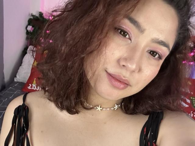 StarlitGaze with curly brown hair, pink eyeshadow, and a subtle smile, wearing a black top and a floral choker, looking directly at the camera. Soft pink lighting.