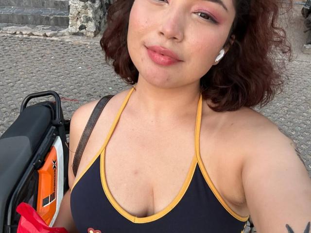 StarlitGaze has curly brown hair, pink eyeshadow, and is wearing a black sports top with yellow trim, showing her tits, holding a bag with an orange strap.