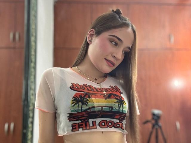 MadisonAngel, con cabello castaño largo, vestida con una camiseta blanca transparente que deja ver pequeños senos, lápiz labial rosa y un collar dorado, de pie en una habitación con un trípode de cámara..