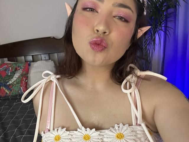 StarlitGaze with pink eyeshadow, puffy lips, and elf ears, blowing kiss, wearing white floral top, exposing small, shiny, wet tits, dark hair, and pink headband in bedroom.