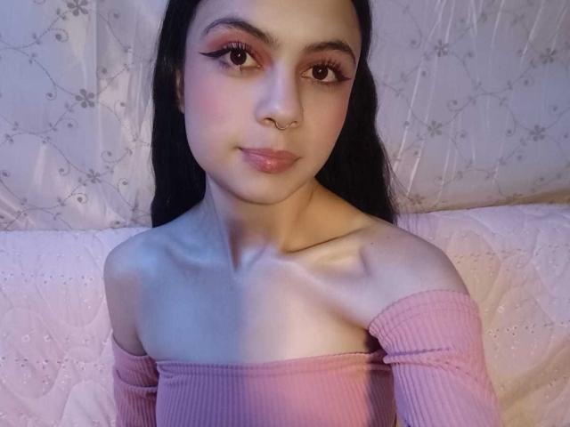 ScarletJhonsonPure with long black hair, pink off-shoulder top, nose ring, and eyeliner, smiling at the camera, showing her bare shoulders and chest, with a subtle pink blush.