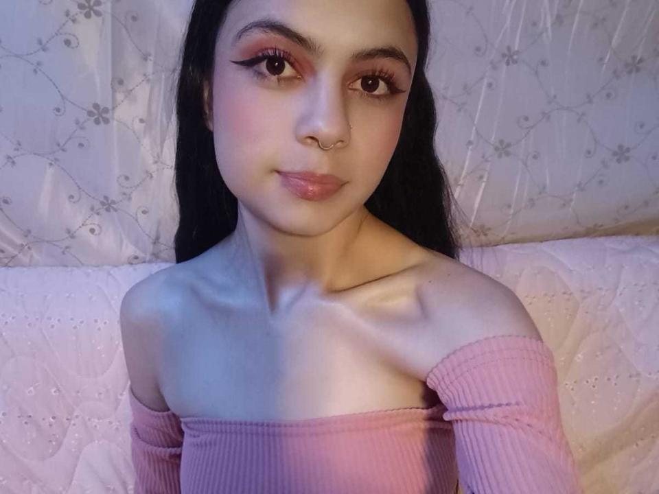 ScarletJhonsonPure with long black hair, pink off-shoulder top, nose ring, and eyeliner, smiling at the camera, showing her bare shoulders and chest, with a subtle pink blush.