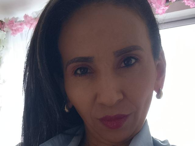 MrsVictoria's face, dark hair, brown eyes, red lipstick, small gold earrings, slight smile, wearing blue shirt, pink eyeshadow, soft light, slightly wrinkled skin, cute, attractive, subtle makeup.