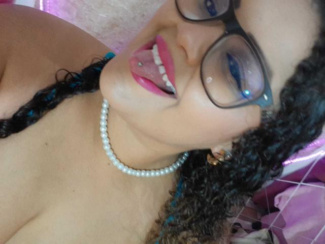 Melannie Carter, con i capelli neri ricci, occhiali, rossetto rosa, la lingua fuori, un piccolo piercing al naso, una collana di perle e con un seno evidente. I suoi glutei non sono visibili..