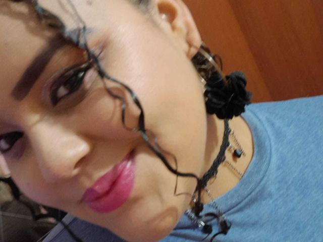MelannieCarter's face with curly black hair, pink lipstick, blue shirt, black earrings, and necklace. Her brown eyebrows are thick and she's giving a slight, playful pout.