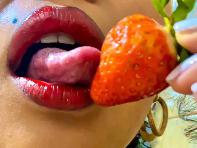 SophieSonyas' glossy red lips and tongue are out, licking a ripe, orange strawberry with tiny seeds. Her wet, shiny lips are slightly parted. She's holding the strawberry with her fingers.