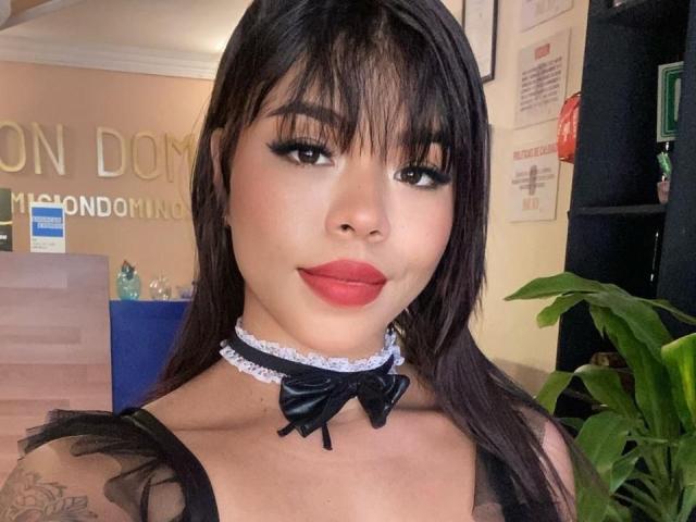 IsisAna mit dunklem Haar, Pony und Eyeliner, trägt roten Lippenstift, eine schwarze Spitzenkette mit Schleife und ein schwarzes Spitzentop. Sie lächelt in die Kamera und zeigt ein kleines Tattoo an der rechten Schulter..