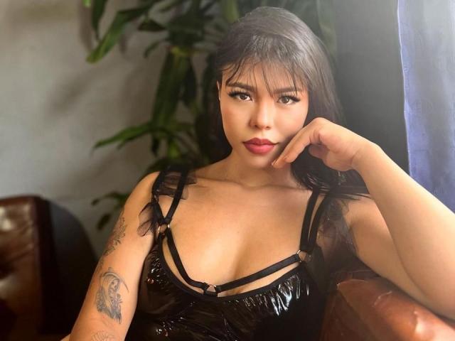 IsisAna with black puffy leather dress, showing her tits, tattoo on right arm, brown hair, red lipstick, hand on cheek, sitting in brown leather chair, green plant behind.