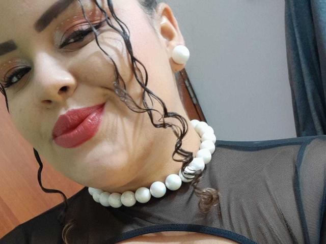 MelannieCarter with curly black hair, wearing red lipstick, black mesh top, white pearl necklace, and white pearl earrings, smiling at the camera. Boobs visible through mesh.