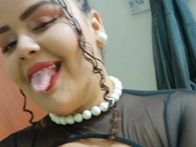 MelannieCarter with curly wet hair, red lipstick, black sheer top, white pearl necklace, and tongue out showing a small blue piercing. Her eyebrows are slightly raised and she's smiling.