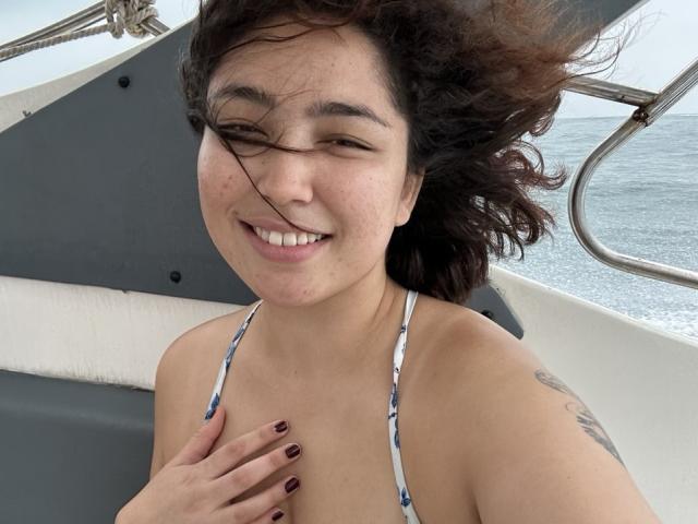 StarlitGaze with curly brown hair, smiling, wearing a blue and white bikini top, hand on chest, nails painted dark red, tattoo on right shoulder, boat in background.