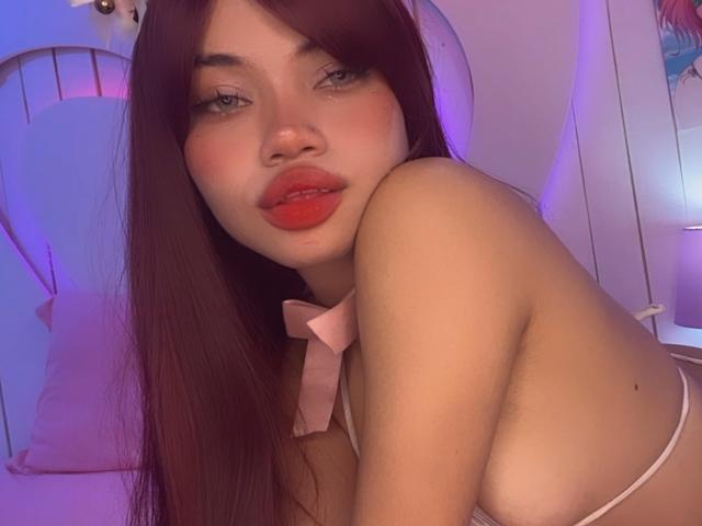 LiliMays with red lipstick, pouty lips, and long brown hair, wearing a pink bow and strappy top, showing her bare shoulders and a hint of her tits.