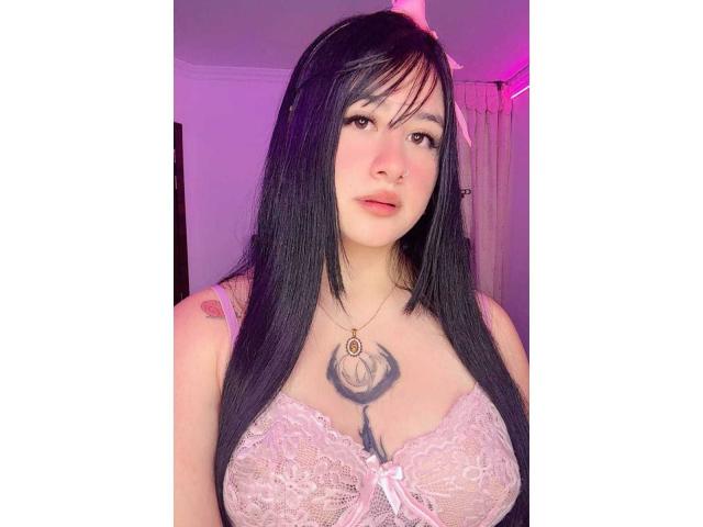 LiliMays has dark red hair, white cat ears with bells, and red lips. She's looking down with eyeliner and a nose piercing, in a purple-lit setting.