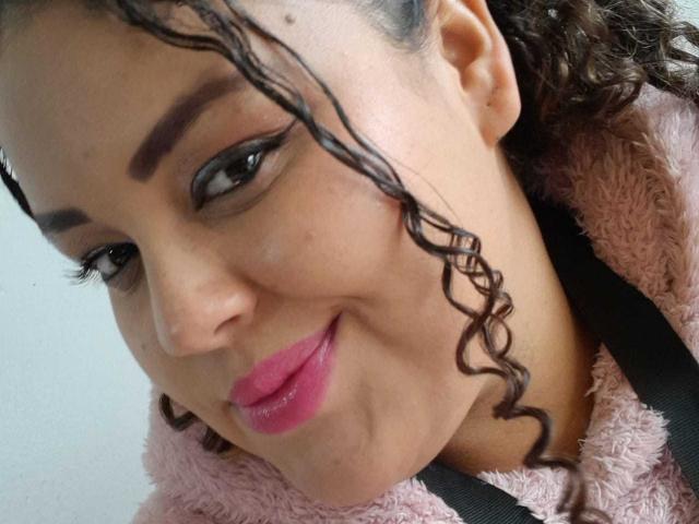MelannieCarter has curly brown hair, pink lipstick, and thick black eyebrows, wearing a pink fuzzy jacket with a black strap, smiling slightly. Her brown eyes are looking to the side.