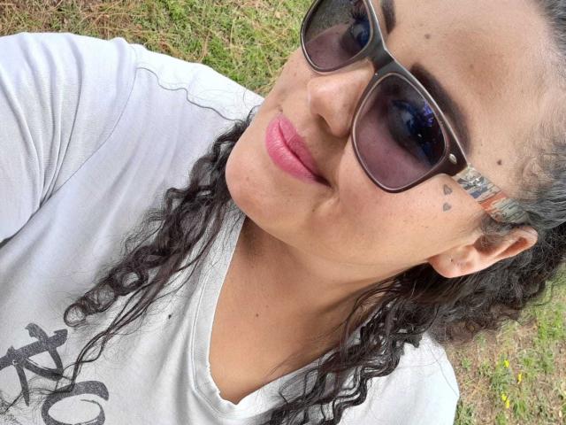 MelannieCarter with curly black hair, wearing sunglasses, white shirt, pink lipstick, and small heart tattoos near her right eye, lying on grass. Her pouty lips are slightly parted.