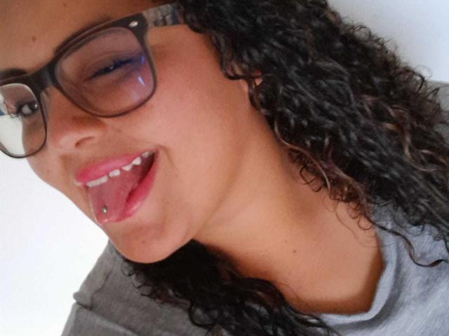 MelannieCarter with curly black hair, wearing glasses, has pink lipstick and a tongue piercing, her tongue out, in a gray shirt, her face slightly tilted.