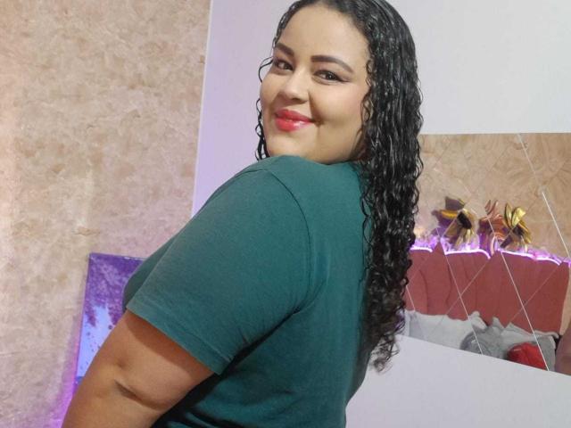 MelannieCarter, curvy Latina woman with long curly black hair, green t-shirt, smiling at camera, red lipstick, standing with back to viewer, looking over shoulder.
