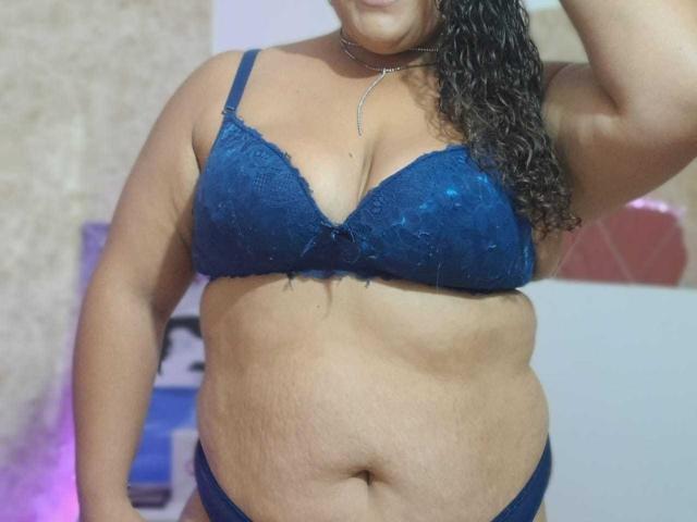 Melannie Carter, con sobrepeso y cabello rizado oscuro, vestida con un sostén y tanga azules de encaje, mostrando sus grandes senos, su abdomen suave y un collar en el cuello, en una foto ligeramente borrosa..