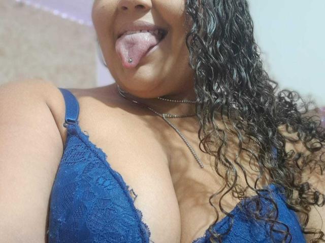 Melannie Carter, com cabelo castanho encaracolado, usando um sutiã de renda azul, com a língua para fora, mostrando seus seios grandes, vestindo um colar de prata, sorrindo e tirando uma selfie..
