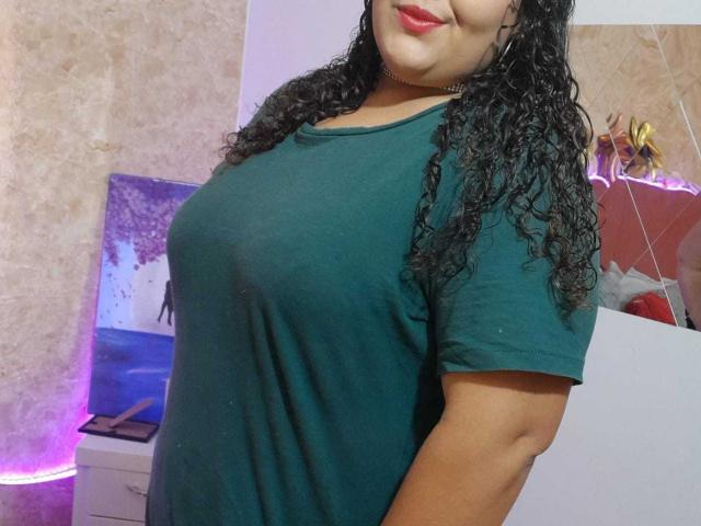 MelannieCarter with curly black hair, wearing a green tee, showing her tits, red lipstick, and a black choker, standing in a room with purple lights.