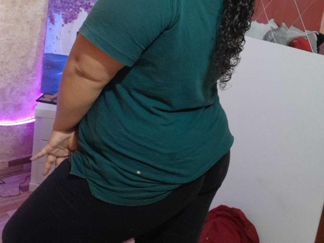 MelannieCarter has a curvy, dark-skinned body with large ass, wearing a green tee and black pants, her curly black hair falling over her back. Her hand's on her butt.