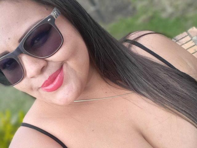 MelannieCarter with black sunglasses, red lipstick, and long straight hair, smiling at the camera, wearing a black spaghetti strap top, showing her boobs.