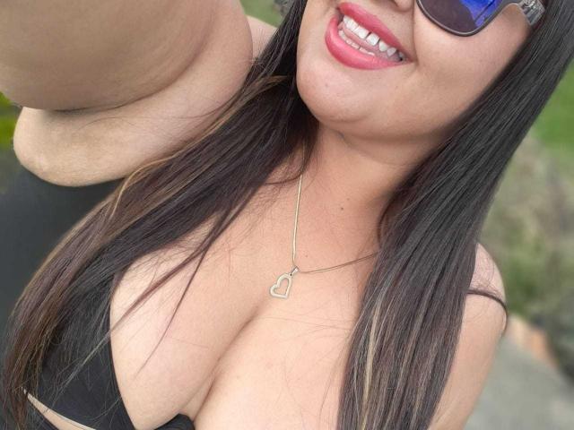 MelannieCarter with long black hair, wearing sunglasses, smiling with pink lipstick, black bikini top showing her big tits, heart necklace, arm raised, selfie, outdoor, sunny.