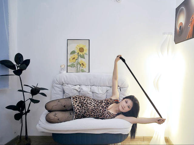 AilsaJade lies on a white couch, wearing a leopard print dress, black fishnet stockings, and holding a black whip, smiling, with long black hair, small tits, and a cute face.