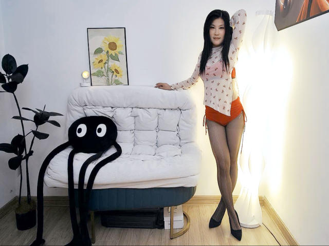 AilsaJade with long black hair, pale skin, wearing a sheer floral top, orange shorts, and black heels, stands by a white tufted couch, with an octopus pillow.