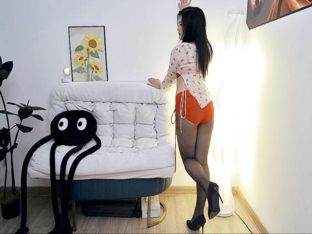 AilsaJade with black hair, wearing a floral shirt, red shorts, and black high heels, stands by a white tufted sofa, touching the wall. Octopus pillow in foreground.
