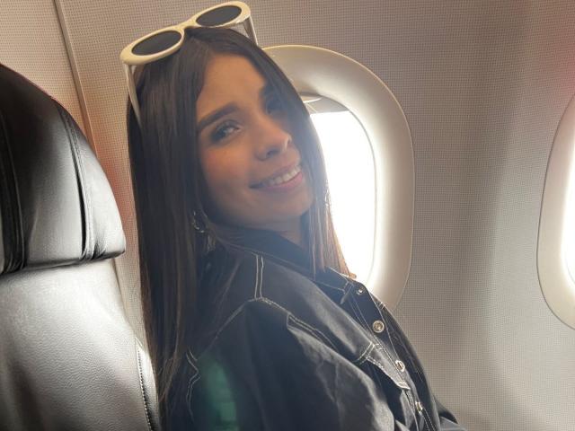 AdalineBliss69 with long brown hair, smiling, wearing a black denim jacket, white-rimmed sunglasses on her head, sitting in an airplane seat, showing her cute ass.