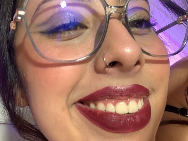 ScarlettParchMusa's face, showing her nose pierced with a small hoop, wearing glasses, with blue eyeshadow, glossy red lipstick, and a slight smile, revealing her teeth.