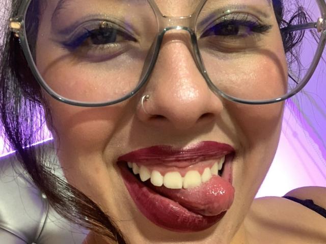ScarlettParchMusa with glasses, nose ring, and red lipstick, tongue out, showing teeth, brown eyes, and dark hair, in a selfie, her face up close, looking playful.