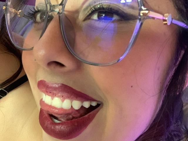 ScarlettParchMusa's face with glossy red lips, white teeth, and purple eyeshadow, wearing clear glasses, looking up with blue reflections in her eyes. Her nose is slightly upturned.