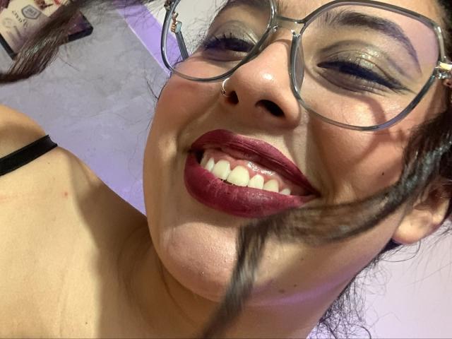 ScarlettParchMusa with big, round glasses, dark lipstick, and shiny eyeshadow, smiling with teeth showing, nose ring, black strap on shoulder, and light brown skin.