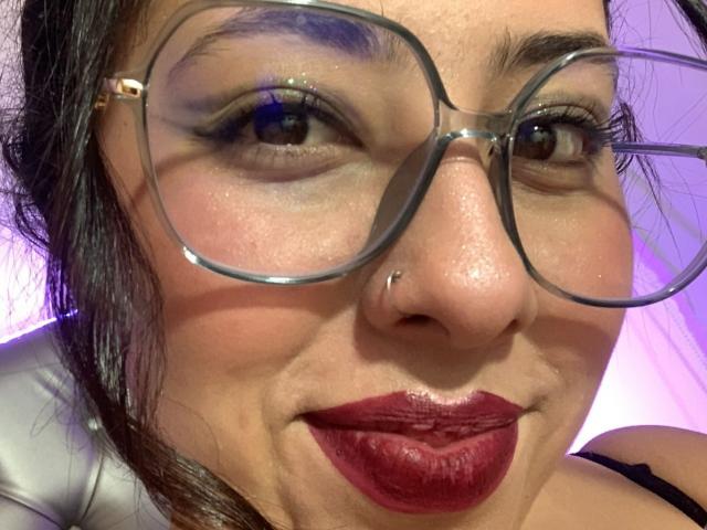 ScarlettParchMusa with glasses, nose piercing, and dark red lipstick, showing a slightly mischievous smile. Her skin is light brown, and she has dark eyebrows and long black hair.