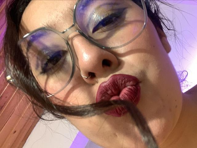 ScarlettParchMusa with big glasses, dark purple lipstick, and a brown hair strand in her pouty lips, blue eyeshadow, nose ring, and a slight purple light in the background.