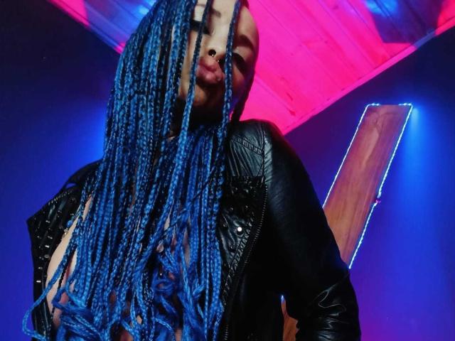 Dalila Musa mit blauen Zöpfen, einer schwarzen Lederjacke und freiliegenden Brüsten, blickt nach unten und trägt einen Nasenring, in einem farbenfrohen, neonbeleuchteten Raum. Ihr Gesicht ist teilweise von ihren Haaren verdeckt..