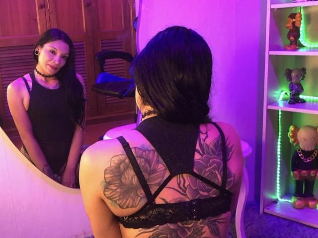 JasminHot69 with black hair, wearing a black lace bra, back tattoo of large flowers, sitting in front of a mirror, reflecting her face, in a purple-lit room.