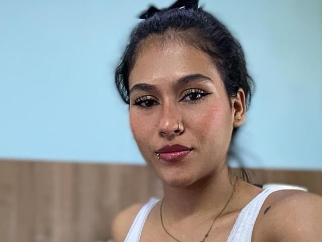 AnnieDarling with dark hair in a ponytail, wearing a white tank top, has a nose ring, lip piercing, and a gold necklace, with a slight smile and visible tattoos.