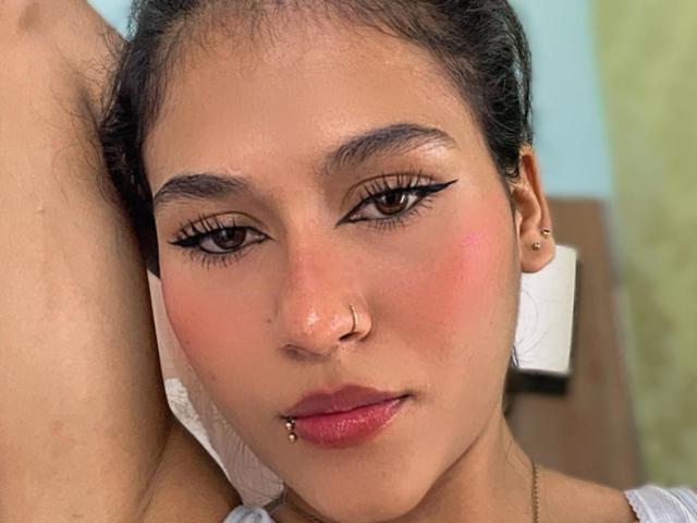 AnnieDarling has medium brown skin, dark hair, and a nose ring. She's wearing bold eyeliner, a lip piercing, and a gold necklace. Her pink cheeks are slightly flushed.