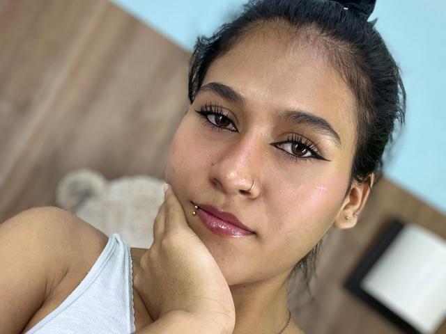 AnnieDarling has dark hair in a bun, brown eyes, and a nose ring. She's touching her chin with her hand, wearing a white tank top, and has glossy lips.