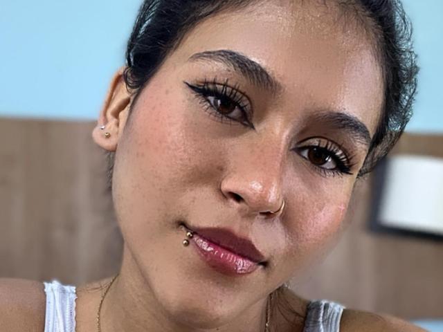 AnnieDarling has brown eyes, black eyeliner, a nose ring, and a lip piercing with a small ball on the bottom. She's wearing a white tank top.