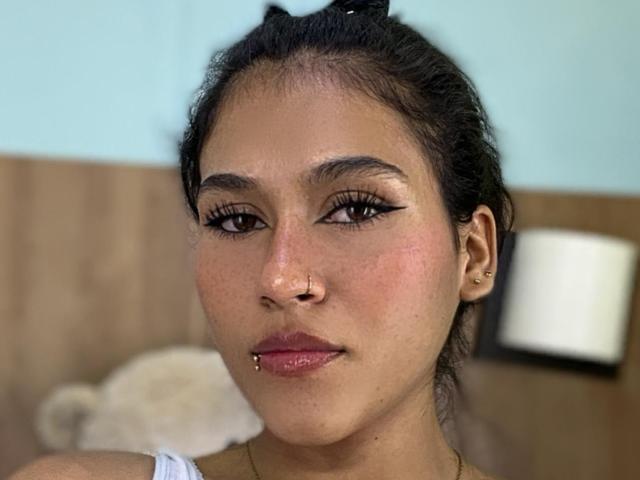 AnnieDarling has a cute face with dark eyes, winged eyeliner, a nose ring, a lip piercing, and a subtle tan. Her hair is up in a small bun.
