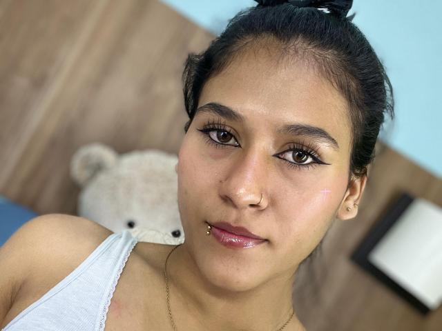 AnnieDarling has tan skin, black hair in a bun, brown eyes, winged eyeliner, nose piercing, lip piercing, white tank top, and a cute bear plushie in the blurry background.
