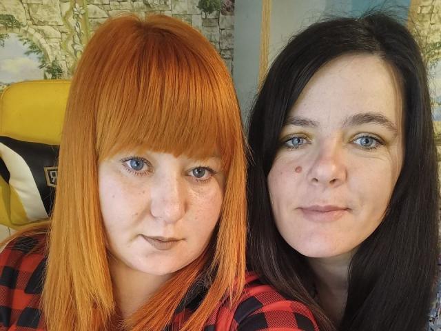 CattieAndSweetie con capelli arancioni e neri, CattieAndSweetie con capelli neri, entrambe che mostrano i loro seni e le lentiggini, in piedi una accanto all'altra, che indossano camicie a quadri rosse e nere..