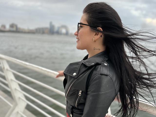 AmelieMuller with black hair, glasses, black leather jacket, and tattoo, standing by a railing, windy, water, city skyline in background. Boobs not visible. Ass not visible. Pussy not visible.