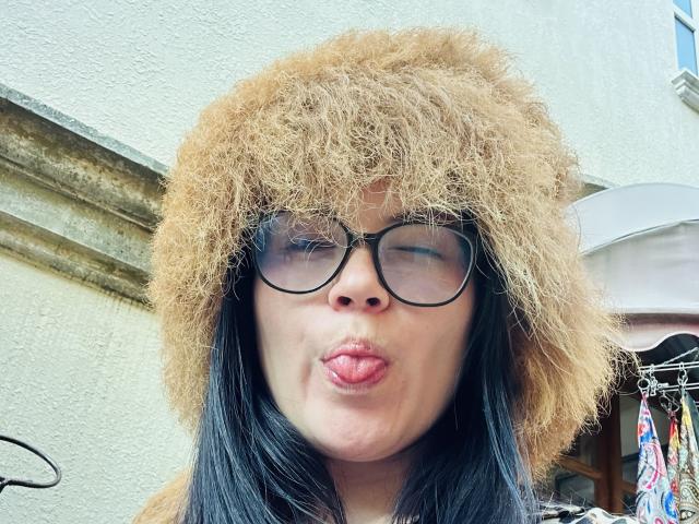 AmelieMuller with black hair, puffy tan fur hat, and round black glasses, puckering her lips in a playful kiss. Her nose is slightly upturned, and she's outside.